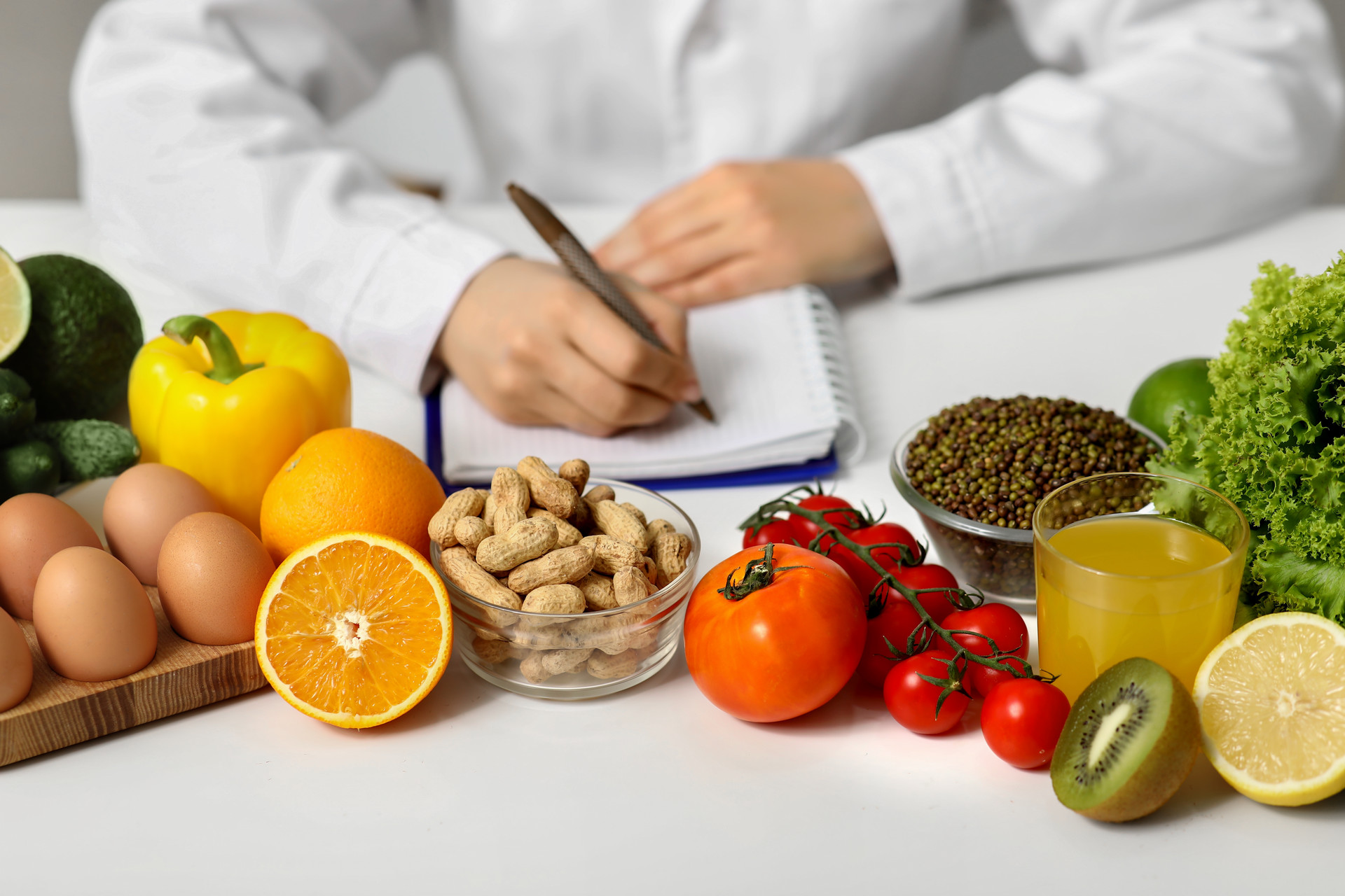 Closeup of nutritionist with healthy food (shelled peanuts, tomatoes, kiwis, eggs, oranges, bell peppers, etc.) writing diet plan in office